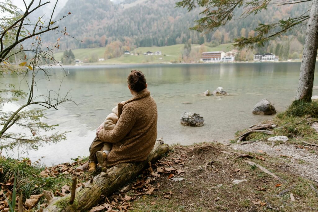 pexels-photo-3933432-3933432 Back view of unrecognizable mother in casual warm outerwear sitting with baby on hands on log near lake and admiring calm scenery in overcast weather